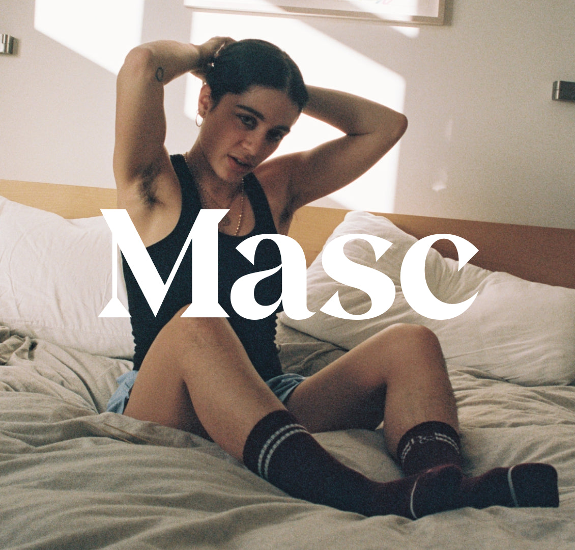 Masc style show through 'Masc' text over photo of woman wearing black tank, sleep shorts and socks, tying their hair back in bed